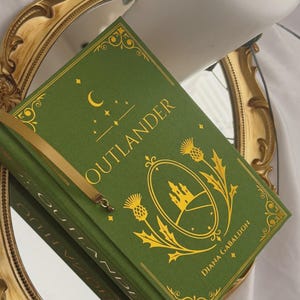 May include: A green hardback book titled Outlander by Diana Gabaldon with gold foil detailing on the cover. The design features a central oval frame containing a castle illustration, flanked by thistle motifs, a crescent moon, and stars.