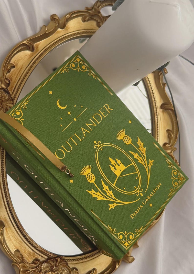 May include: A green hardback book titled Outlander by Diana Gabaldon with gold foil detailing on the cover. The design features a central oval frame containing a castle illustration, flanked by thistle motifs, a crescent moon, and stars.