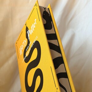 May include: A hardback book titled Harry Potter and the Chamber of Secrets with a bright yellow cover featuring a large black snake illustration. The page edges are decorated with a black and white snake pattern that continues along the side.