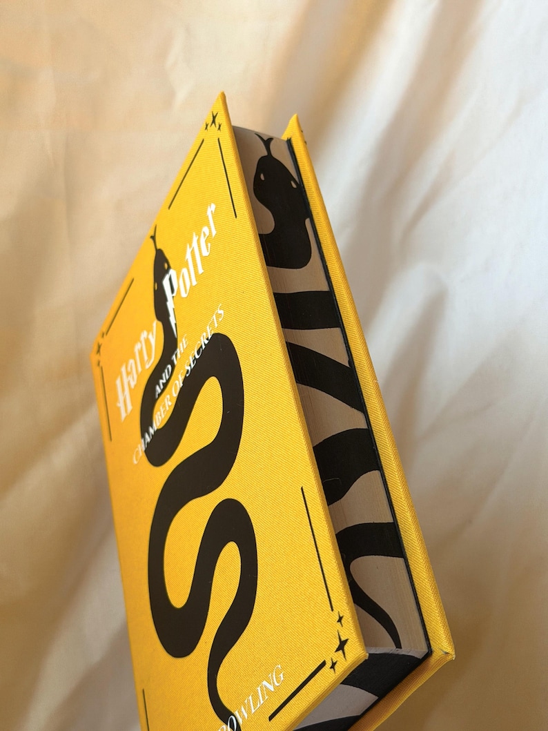 May include: A hardback book titled Harry Potter and the Chamber of Secrets with a bright yellow cover featuring a large black snake illustration. The page edges are decorated with a black and white snake pattern that continues along the side.