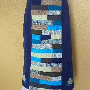 May include: A navy blue wrap skirt with a patchwork design. The patchwork features rectangular panels in shades of blue, green, and brown with paisley and floral patterns. The skirt has a white ruffled hem and embroidered floral accents.