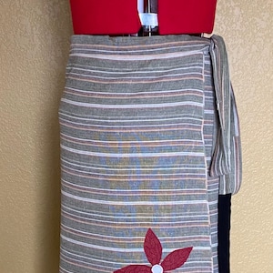 May include: A wrap skirt with horizontal stripes in shades of gray, brown, and white, adorned with appliqued flower designs in black, red, green, and beige. The skirt is displayed on a red dress form.