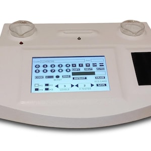 May include: A white electronic diagnostic device featuring a central touchscreen interface with numerical and function buttons, including labels like Code, Copy, Neut, Auto, Diag, Imprint, Erase, and Save. Two clear glass beakers sit on the top surface.