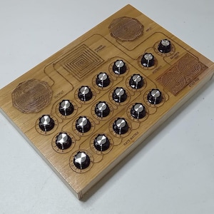 May include: A rectangular wooden device featuring a grid of fifteen black and silver control knobs. The surface is engraved with labels including Output, Power Pyramid, Input, Balance Test, Potency, Stick Pad, and Rate Matrix with circular patterns.