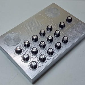 May include: A rectangular silver electronic device featuring a grid of sixteen black control knobs with silver tops. The surface includes etched circular and square patterns, with text labels including Rate Matrix, Output, and Potency printed in black.