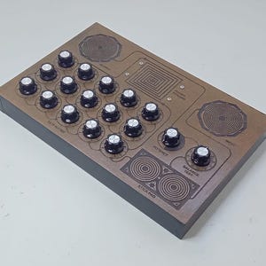 May include: A rectangular electronic device with a brown top panel featuring numerous black control knobs with silver centers. The panel includes engraved labels such as Rate Matrix, Output, Power Pyramid, Input, Potency, Balance Test, and Stick Pad.
