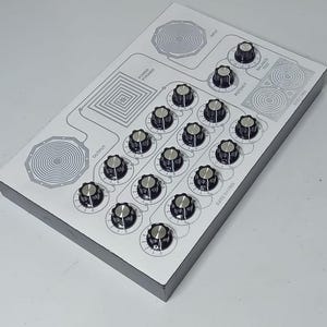 Pode incluir: Um dispositivo eletrônico retangular com um painel superior branco apresentando botões de controle pretos e prateados, padrões gráficos circulares e quadrados, e etiquetas incluindo Input, Output, Power Pyramid, Balance Test, Potency, Stick Pad e Rate Matrix.
