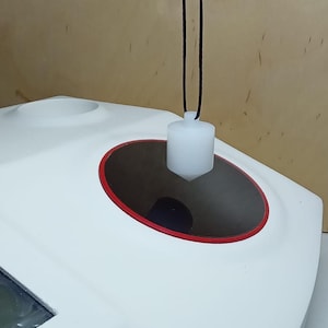 May include: A white cylindrical plumb bob with a pointed tip suspended by a thin dark cord over a white surface featuring a circular opening with a red rim. The object is positioned above a dark, recessed circular area within the white housing.