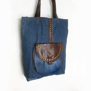 Könnte beinhalten: Eine blaue Jeans-Tragetasche mit einer braunen Ledertasche mit Klappe, die mit goldenen Nieten verziert ist. Die Tasche hat braune Lederriemen.