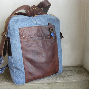 Könnte beinhalten: Eine blaue Jeans- und braune Leder-Messenger-Tasche mit einem langen verstellbaren Riemen. Die Tasche hat eine große Tasche auf der Vorderseite mit einem Reißverschluss.