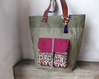 May include: A green canvas tote bag with a pink and patterned pocket and a brown leather strap. The bag has a blue tassel hanging from it.