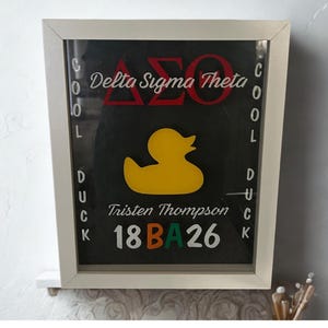 Może przedstawiać: Czarny, oprawiony znak z białą obwódką. Znak zawiera czerwony tekst "Delta Sigma Theta", żółtą grafikę kaczki oraz imię "Tristen Thompson" z numerami "18BA26". Słowa "COOL DUCK" są napisane pionowo po bokach.