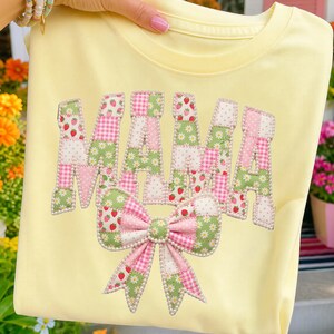 May include: A pale yellow t-shirt featuring the word MAMA in large, block letters and a bow graphic below. The letters and bow are filled with a patchwork pattern of strawberries, green daisies, pink gingham, and white polka dots, outlined in white beads.