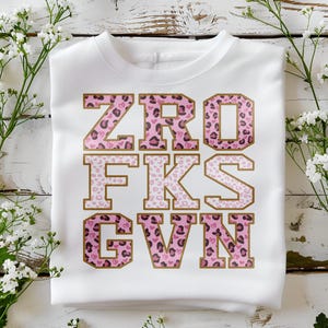 Puede incluir: Sudadera blanca de cuello redondo con el texto "ZRO FKG GVN" en un diseño de estampado de leopardo rosa con ribete dorado. Las letras están en una fuente de estilo universitario. La sudadera se muestra sobre una superficie de madera blanca con flores blancas.