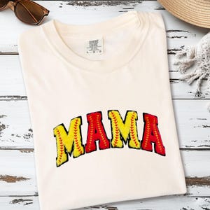 May include: Cream-colored t-shirt with the word "MAMA" in a baseball-themed design. The letters are yellow and red, with black outlines and stitching details. The shirt is folded on a light wood surface.