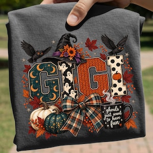 Può includere: Una t-shirt grigio scuro con il nome GIGI in grandi lettere decorate. Il design include un cappello da strega, due gufi neri, foglie autunnali, zucche, un fiocco scozzese e una tazza nera con il testo ghouls just want to have fun.