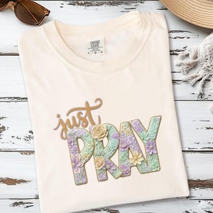 Può includere: T-shirt color crema con le parole "just PRAY" in oro e disegni floreali. La parola "PRAY" è decorata con fiori dai colori pastello. La maglietta è piegata su una superficie di legno bianco.