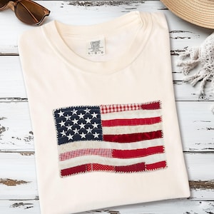 Puede incluir: Camiseta color crema con un diseño de bandera estadounidense en patchwork. La bandera incorpora rayas rojas y blancas, un campo azul con estrellas blancas y varios patrones de tela. La camiseta está doblada sobre una superficie de madera blanca.