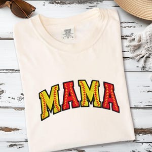 May include: Cream-colored t-shirt with the word "MAMA" in a baseball-themed design. The letters are yellow with red stitching, outlined in black. The shirt is folded on a white wooden surface.