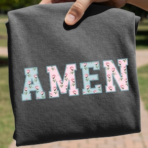 Könnte beinhalten: Ein dunkel anthrazitfarbenes T-Shirt mit dem Wort AMEN in großen Blockbuchstaben. Die Buchstaben haben einen hellrosa Hintergrund mit einem Muster aus kleinen roten Rosen und grünen Blättern, umrandet von einer hellblauen gepunkteten Linie.
