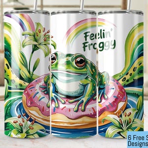 May include: A white tumbler featuring a green frog perched on a pink and white doughnut. The background is a swirling green and yellow design with the text "Feelin' Froggy".