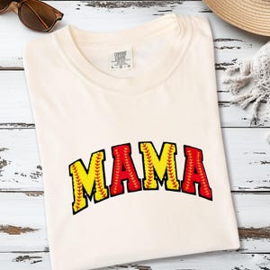 May include: Cream-coloured t-shirt featuring the word "MAMA" in a baseball-themed design. The letters are yellow with red stitching and a red outline, resembling a baseball. The shirt is folded on a light wood surface.