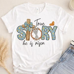 May include: A white short-sleeve t-shirt featuring a graphic print with the text True Story he is risen. The design includes a wooden cross with a blue floral bow, a crown of thorns with three nails, and an orange butterfly on a blue floral background.