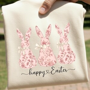May include: A cream-colored shirt featuring a graphic of three bunnies with a pink floral pattern. Each bunny has a small cream-colored bow around its neck. Below the bunnies, the text "happy Easter" is written in a black cursive script font.