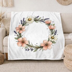 May include: A white throw blanket featuring a central circular wreath illustration composed of watercolor-style pink blossoms, white daisies, and purple lavender sprigs with green leaves, arranged in a delicate botanical pattern on the fabric.