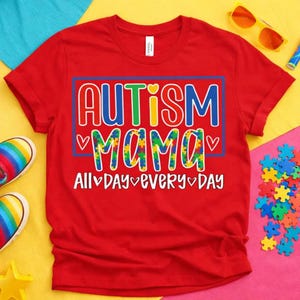 Puede incluir: Una camiseta roja de manga corta con el texto "AUTISM MAMA" en letras coloridas con patrón de rompecabezas dentro de un borde rectangular azul, con el texto "ALL DAY EVERY DAY" debajo, acentuado por pequeños iconos de corazones blancos.
