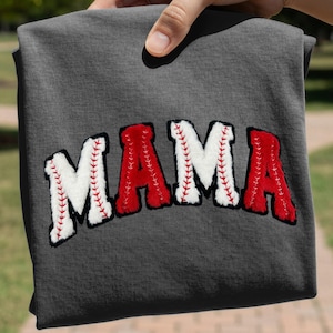 May include: Dark gray t-shirt with the word "MAMA" in a baseball-themed design. The letters are red and white, with black outlines and red stitching, resembling baseballs. The shirt is held up by a hand.