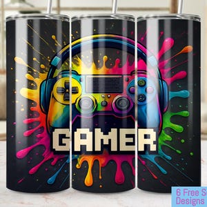 Puede incluir: Un vaso negro con un diseño de salpicaduras de pintura de colores y un gráfico de un mando de videojuego. El texto "GAMER" está en letras pixeladas.