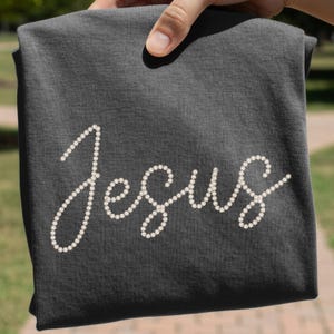 May include: Dark gray t-shirt with the word "Jesus" spelled out in white pearl-like embellishments. The shirt is held up by a hand, showcasing the design. The fabric appears soft and comfortable.