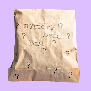 Op de afbeelding: Een bruine papieren zak met de tekst "mystery/? bead? bag?" erop gedrukt. De zak is gevuld met vraagtekens.