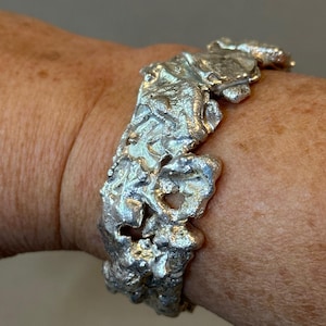 May include: A silver bracelet with a textured, organic design. The bracelet is made of a single piece of metal and has a rough, irregular shape.