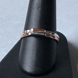 May include: Two thin hammered metal rings, one silver and one rose gold, stacked on a ring mandrel.