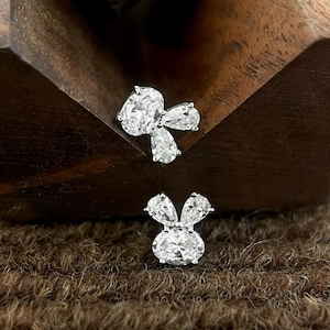 May include: Two silver stud earrings. One is a bow shape with an oval centre stone and three pear-shaped stones. The other is a bunny shape with an oval centre stone and two pear-shaped stones for the ears. The earrings are on a brown surface.
