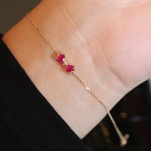 May include: A delicate gold bracelet featuring a bow design with two heart-shaped, ruby-colored gemstones. A small diamond accent is set between the hearts. The bracelet is shown on a wrist.