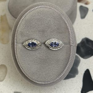 May include: A pair of silver earrings featuring a blue gemstone surrounded by smaller white gemstones. The earrings are displayed in a velvet box.