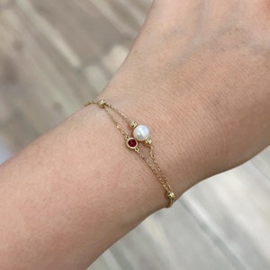 May include: A gold chain bracelet featuring a white pearl and a red gemstone. The bracelet is layered with two chains.