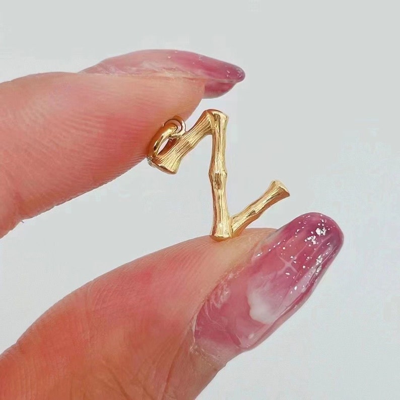 May include: Gold-toned letter Z charm with a bamboo-like design. The charm is held in a hand with pink nail polish.