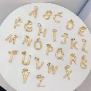 May include: A set of 26 gold-coloured alphabet letter charms. Each letter is made of a textured metal that resembles bamboo. The charms are arranged in alphabetical order on a white surface.