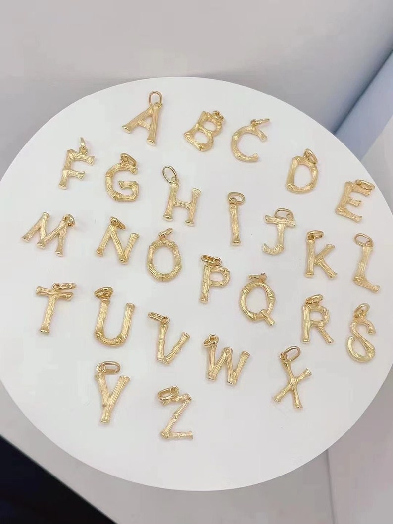 May include: A set of 26 gold-coloured alphabet letter charms. Each letter is made of a textured metal that resembles bamboo. The charms are arranged in alphabetical order on a white surface.