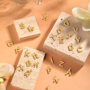 May include: A set of gold-toned alphabet letter charms. The charms are made of a textured metal and are arranged on a light brown surface.