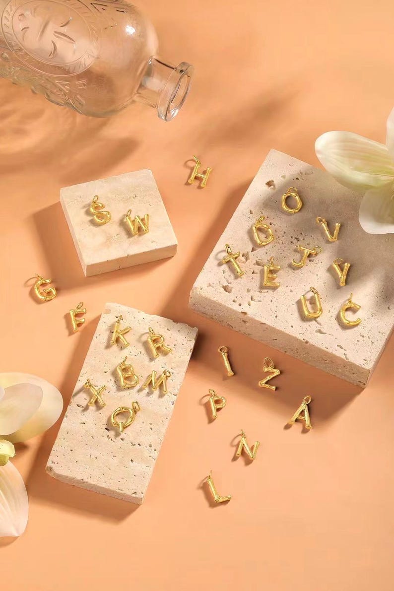 May include: A set of gold-toned alphabet letter charms. The charms are made of a textured metal and are arranged on a light brown surface.