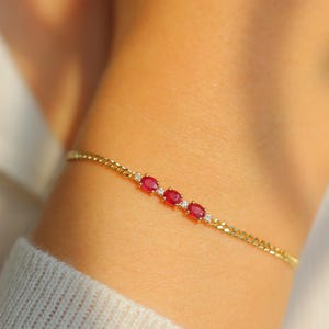 Könnte beinhalten: Ein goldenes Kettenarmband mit drei roten ovalen Edelsteinen und kleinen Diamanten auf jeder Seite der Edelsteine.