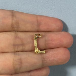 May include: Gold-toned charm in the shape of the letter L. The charm has a textured, wood-like finish.