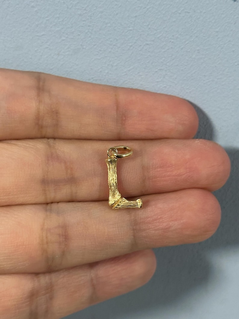 May include: Gold-toned charm in the shape of the letter L. The charm has a textured, wood-like finish.