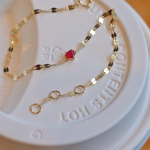 May include: A gold chain bracelet with a small red heart charm. The bracelet is on a white coffee cup lid with the text "Contents Hot" printed on it.