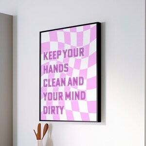 Pode incluir: Uma impressão de arte de parede emoldurada com uma borda preta. A impressão apresenta um fundo xadrez rosa e branco com o texto "KEEP YOUR HANDS CLEAN AND YOUR MIND DIRTY" em uma fonte roxa em negrito. Um vaso branco, uma tigela e um recipiente cor de cobre estão na bancada.